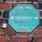 Porcelain house beside Liverpool Irish Famine plaque on red brick wall.