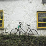 A dilapidated bicycle leans on a cottage wall with bright yellow painted sash windows